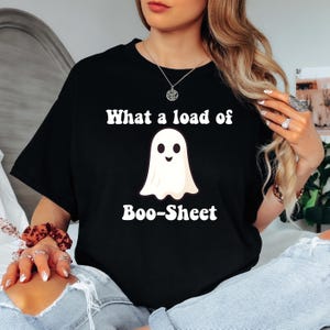 Puede incluir: Camiseta negra con el texto "What a load of Boo-Sheet" sobre un gráfico de fantasma sonriente. El fantasma es blanco con una cara sonriente negra. La camiseta es de estilo casual.
