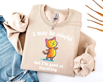 May include: Beige sweatshirt with the text "I may be stupid but I'm good at kayaking" and a cartoon cat illustration. The cat is orange and yellow with a green base. The sweatshirt is paired with blue jeans and a necklace.