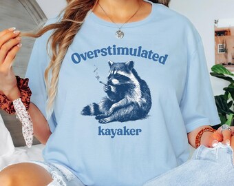 May include: Light blue t-shirt with the words "Overstimulated kayaker" in dark blue. The shirt features a detailed illustration of a raccoon smoking a cigarette. The raccoon is sitting down, and the design is centered on the shirt.