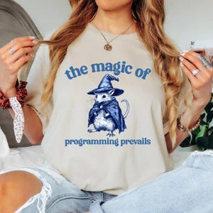 Op de afbeelding: Een beige t-shirt met een blauwe afbeelding van een muis met een tovenaarshoed en een cape. De tekst "the magic of programming prevails" is in blauw onder de afbeelding gedrukt.