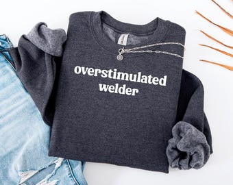 May include: A charcoal grey sweatshirt with the words "overstimulated welder" printed in white. The sleeves are cuffed. A silver necklace with a pendant rests on the sweatshirt. Faded blue jeans are also visible.