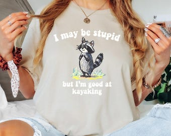 May include: Beige t-shirt with a raccoon graphic and the text "I may be stupid but I'm good at kayaking". The raccoon is depicted in black, gray, and white, standing on green grass. The shirt is a casual, comfortable style.