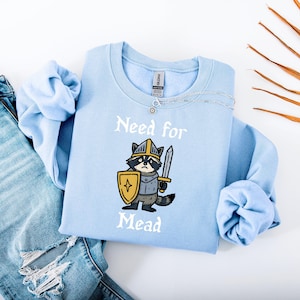 Op de afbeelding: Een lichtblauw sweatshirt met een cartoon wasbeer ridder met een zwaard en schild. De tekst "Need for Mead" is in het wit gedrukt. Het sweatshirt wordt getoond met een blauwe jeans en een zilveren ketting.