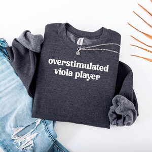 Puede incluir: Sudadera gris oscuro con el texto "overstimulated viola player" en blanco. Un collar plateado con un colgante está sobre la camisa. La sudadera está doblada junto a unos vaqueros azul claro.