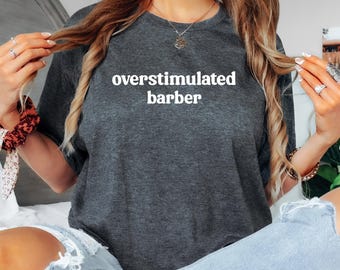 Może przedstawiać: Ciemnoszara koszulka z białym napisem "overstimulated barber". Koszulka ma okrągły dekolt i wydaje się być wykonana z miękkiego materiału. Tekst jest wyśrodkowany na piersi.
