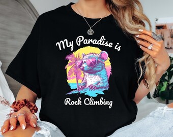 Può includere: Maglietta nera con una grafica di un procione che indossa occhiali da sole e tiene un albero di palma. Il testo "My Paradise is Rock Climbing" è stampato sotto il procione.