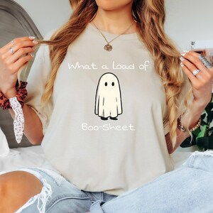 Puede incluir: Camiseta beige con la frase "What a load of Boo-Sheet" y una ilustración de un fantasma de dibujos animados. El fantasma es blanco con ojos y pies negros. La camiseta es de estilo casual.
