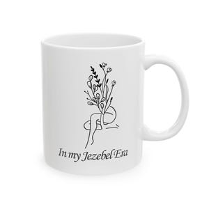 May include: White ceramic mug with a black line drawing of a person with flowers growing out of their head. The text "In my Jezebel Era" is printed on the mug.