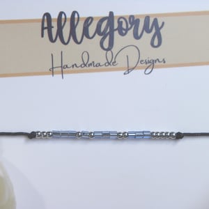 Op de afbeelding: Een zwarte koordarmband met een reeks kleine metallic zilveren bolletjes en lichtblauwe cilindervormige kralen. De armband wordt gepresenteerd op een witte kaart met de tekst Allegory Handmade Designs in zwarte letters gedrukt.