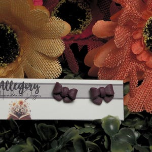 May include: A pair of dark purple bow-shaped stud earrings displayed on a white rectangular card. The card features the text Allegory Handmade Designs, a small illustration of a bouquet of flowers, and a QR code in the bottom left corner.