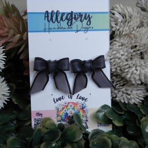 May include: A pair of dark grey bow-shaped stud earrings attached to a white display card. The card features the text Allegory Handmade Designs at the top and Love is Love with a colourful floral and rainbow graphic at the bottom centre.