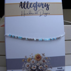 Può includere: Un braccialetto delicato con perline alternate azzurre e argento metallizzato su un sottile cordino bianco, esposto su una carta bianca con l'etichetta Allegory Handmade Designs, una grafica floreale e un piccolo logo Venmo.