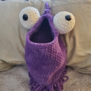 May include: Purple crocheted toy with large white eyes and black pupils. The toy has a long, fringed bottom and a large opening at the top.