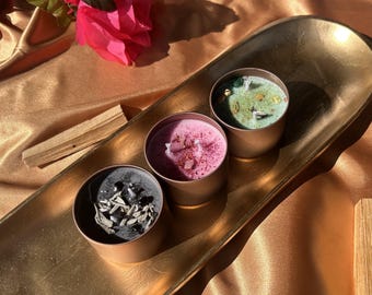 May include: Three small candles in copper-colored tins sit on a gold-colored tray. One candle is black with white herbs, another is pink with pink crystals, and the third is green with gold flecks. A pink flower and a piece of wood are also in the image.