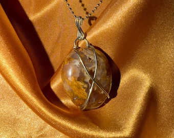 May include: A pendant necklace featuring a polished, oval-shaped stone with golden and brown hues, wrapped in silver-colored wire. The stone hangs from a delicate, gold-colored chain, set against a backdrop of golden fabric.