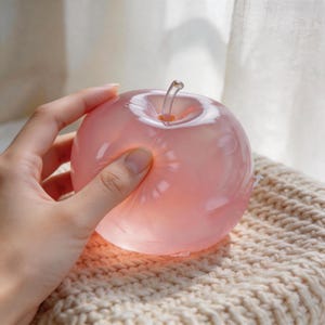 May include: A translucent pink glass apple with a clear stem. The apple is being held by a hand, and the light shines through the glass. The apple is sitting on a beige knitted surface.