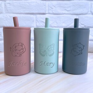 May include: Three personalized silicone cups with straws. The cups are pink, light blue, and gray. The pink cup has a lion design and the name "Jackie" engraved on it. The light blue cup has a butterfly design and the name "Mary" engraved on it. The gray cup has a fish design and the name "Jackie" engraved on it.