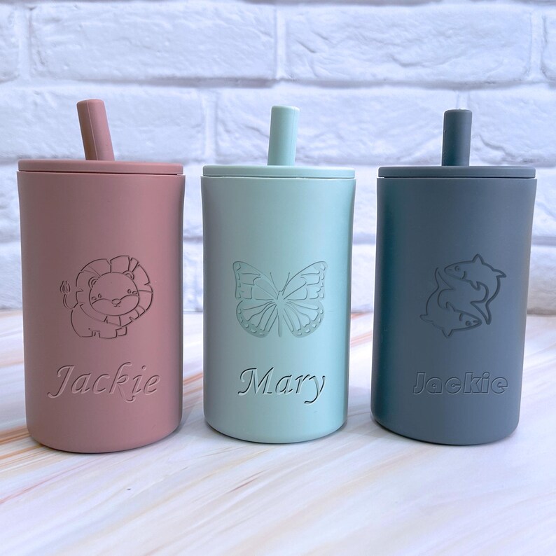 May include: Three personalized silicone cups with straws. The cups are pink, light blue, and gray. The pink cup has a lion design and the name "Jackie" engraved on it. The light blue cup has a butterfly design and the name "Mary" engraved on it. The gray cup has a fish design and the name "Jackie" engraved on it.