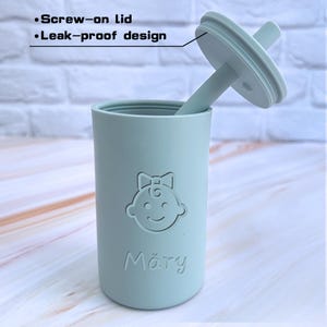 May include: A light blue silicone cup with a screw-on lid and a straw. The cup has a girl's face and the name "Mary" embossed on the front.