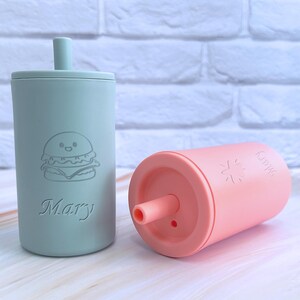 May include: Two personalized silicone cups with straws. The green cup has a cartoon burger and the name "Mary" on it. The pink cup has a sun and the name "Kamrie" on it.