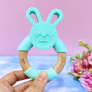 May include: A teal blue silicone bunny-shaped teether attached to a wooden ring. The ring is engraved with the word "Baby".