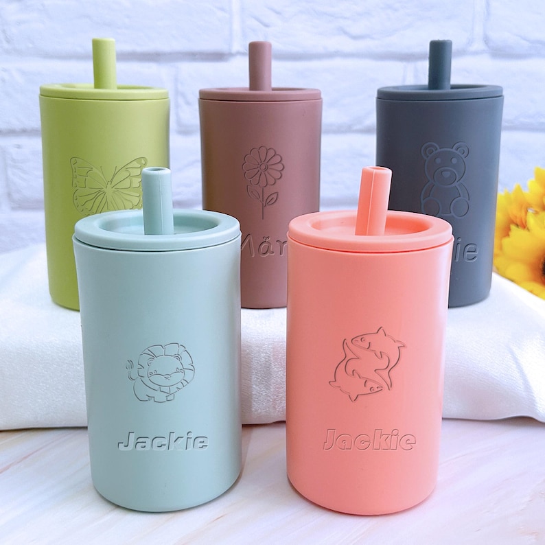 May include: Five colorful silicone cups with built-in straws and different animal designs. The cups are labeled with the names "Jackie" and "M&aring;r", and feature a butterfly, flower, bear, lion, and fish.