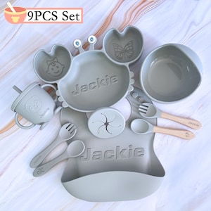 May include: A 9 piece silicone baby feeding set in gray with a crab-shaped plate, a bowl, a cup, a bib, a spoon, a fork, a suction plate, and two spoons. The set is personalized with the name "Jackie".