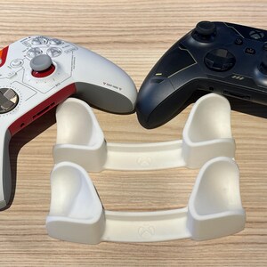 Könnte beinhalten: Zwei Videospiel-Controller, einer weiß mit roten Akzenten und der andere schwarz, werden mit einem weißen Controller-Ständer gezeigt. Der weiße Controller hat ein farbenfrohes Design, während der schwarze einen schlanken, modernen Look hat.