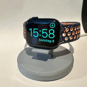 Könnte beinhalten: Eine schwarze Apple Watch mit einem bunten Musterband befindet sich auf einem grauen Ladeständer. Das Zifferblatt der Uhr zeigt die Zeit 15:58 und das Datum "Sonntag 8".