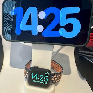 Könnte beinhalten: Ein weißer Ständer mit einer schwarzen und roségoldenen Apple Watch mit einem schwarzen Armband. Das Zifferblatt der Uhr zeigt die Zeit 14:25 und das Datum Donnerstag 22. Ein schwarzes Smartphone mit einem blauen Bildschirm, auf dem die Zeit 14:25 angezeigt wird, befindet sich auf dem Ständer.