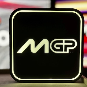 Könnte beinhalten: Ein schwarzes quadratisches Schild mit weißem Rand und dem Text "MGP" in weißen Buchstaben. Das Schild wird von hinten beleuchtet, wodurch ein heller Schein um die Buchstaben entsteht.