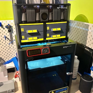 Könnte beinhalten: Ein schwarzer 3D-Drucker mit einer durchsichtigen Tür und einem roten Display. Der Drucker hat einen Aufkleber mit der Aufschrift "Replicator U.S.S. Enterprise". Auf dem Drucker befindet sich ein schwarzes Filament-Lager mit vier Schubladen, die mit 1, 2, 3 und 4 beschriftet sind. Das Lager hat einen durchsichtigen Deckel mit Filamentspulen im Inneren.