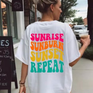Könnte beinhalten: Weißes T-Shirt mit dem Schriftzug "SUNRISE SUNBURN SUNSET REPEAT" in bunten, Retro-Lettern. Das Shirt hat eine lässige, übergroße Passform, ideal für einen Tag am Strand oder einen Sommerurlaub.