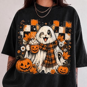 May include: A black t-shirt featuring a graphic of a white cat dressed as a ghost, wearing an orange and black plaid scarf. The cat holds a jack-o'-lantern mug with marshmallows. The design includes orange pumpkins, autumn leaves, and a checkered background.