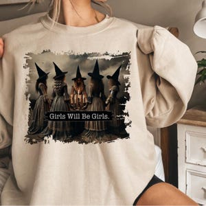 Puede incluir: Una sudadera beige con un estampado gráfico de cinco brujas con sombreros y túnicas negras de pie alrededor de un caldero con velas. El texto "Girls Will Be Girls." está impreso debajo de las brujas.