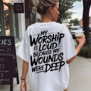 May include: A white t-shirt with the text "MY WORSHIP IS LOUD BECAUSE MY WOUNDS WERE DEEP" in black. The shirt is worn by a person walking on a pavement, holding a beverage.