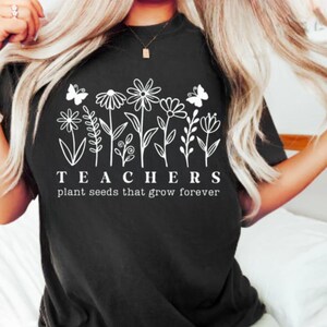 May include: Black t-shirt with a white floral design and the text "TEACHERS plant seeds that grow forever." The design includes various flowers and butterflies. The shirt has a crew neck.
