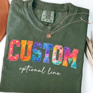 May include: Olive green t-shirt with the word "CUSTOM" in colorful, embroidered letters. The letters are outlined in a rainbow of colors and have a star pattern. The text "optional line" is below the word "CUSTOM". A silver necklace with a pendant is on the shirt.
