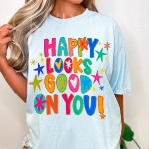 May include: Light blue t-shirt with the text "HAPPY LOOKS GOOD ON YOU!" in colorful, playful lettering. The design includes stars, hearts, and smiley faces. A casual, comfortable tee.