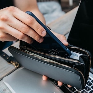 May include: A black leather zip-around wallet with multiple interior compartments, containing a dark blue smartphone and a white card. The wallet features a metal zipper closure and a detachable black leather wrist strap with a gold-tone metal clasp.