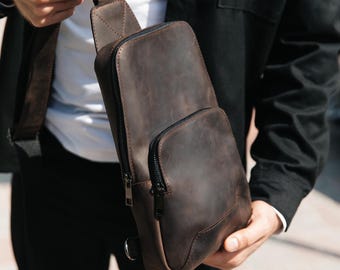 May include: A dark brown leather sling bag featuring a vertical main compartment with a black zipper and a smaller front pocket with a matching black zipper. The bag includes a single adjustable shoulder strap attached to the top and side.