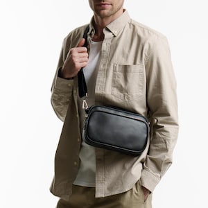 May include: A black rectangular crossbody bag with a black strap and silver hardware. The bag is worn over the shoulder. The person is wearing a tan button-down shirt and a white t-shirt.
