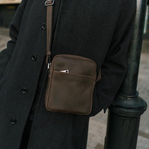 Könnte beinhalten: Eine braune Leder-Umhängetasche mit einer Vordertasche und einem verstellbaren Riemen. Die Tasche wird über einem dunkelgrauen Mantel getragen. Die rechteckige Form und das schlichte Design deuten auf eine minimalistische Ästhetik hin.