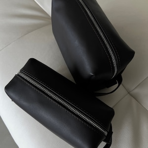 Pode incluir: Duas bolsas de couro preto com zíper. As bolsas são retangulares e têm um zíper prateado. As bolsas estão sobre uma superfície branca.