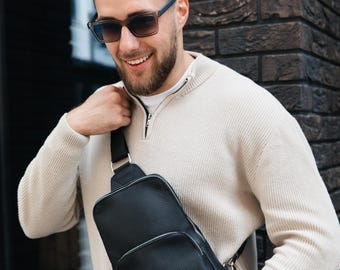 Pode incluir: Uma bolsa tiracolo de couro preto com superfície texturizada, apresentando um compartimento principal com zíper e um bolso frontal menor com zíper. A bolsa é usada com uma alça preta ajustável sobre o peito, combinada com um suéter bege.