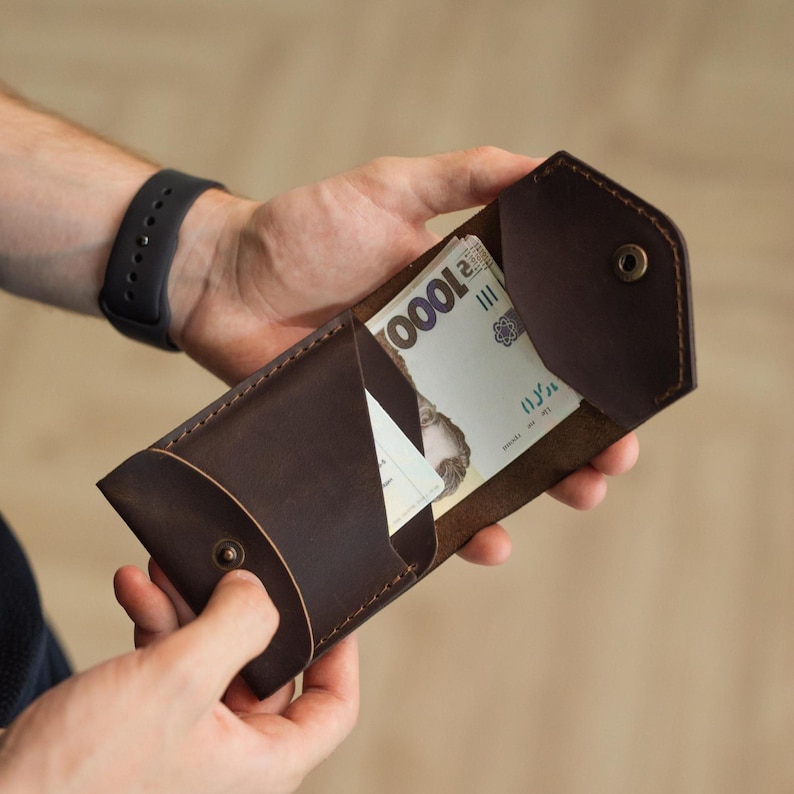May include: A dark brown leather wallet, open to reveal a wad of cash and a card. The wallet is held open, showcasing its interior compartments and the button closure. The leather has a rich, textured appearance.