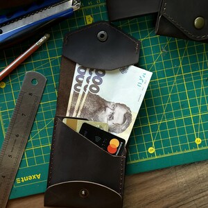 May include: A dark brown leather wallet with a snap closure, holding paper currency and credit cards. The wallet is open, revealing the contents. A ruler, pencil, and utility knife are visible in the background.