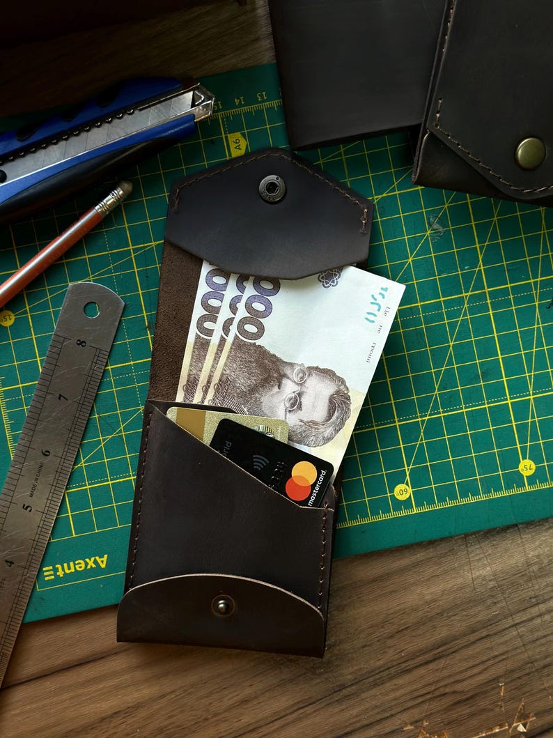 May include: A dark brown leather wallet with a snap closure, holding paper currency and credit cards. The wallet is open, revealing the contents. A ruler, pencil, and utility knife are visible in the background.