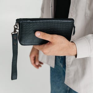 Peut inclure: Un portefeuille rectangulaire noir avec un motif texturé en peau de crocodile. Le portefeuille présente une fermeture éclair sur le dessus et une dragonne en cuir noir assortie fixée sur le côté par un fermoir pivotant en métal sombre.