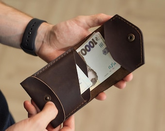 May include: A dark brown leather wallet with a pointed flap and a metal snap closure. The wallet contains a 1000 denomination banknote and a white card. The leather features visible stitching along the edges and a matte, textured finish.